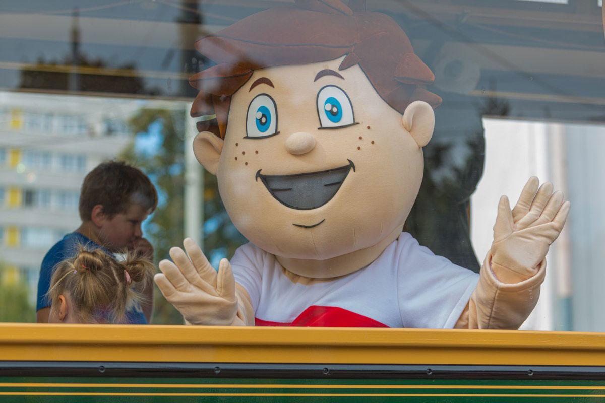 Na fotografii widnieje maskotka w postaci kostiumu chłopca uchwycona przez szybę autobusu